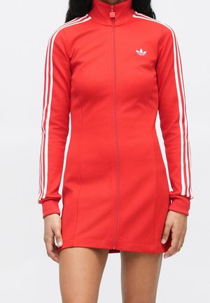 Kobieta w czerwonej sukience na zamek marki Adidas z białymi trzema paskami na rękawach i małym logo na piersi na białym tle.