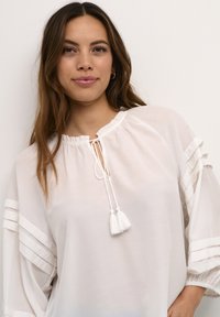 Blusa bianca e leggera con scollatura arricciata, cordoncino con nappine e dettagli arricciati su maniche e spalle.