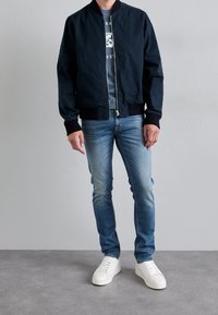 Chaqueta bomber azul marino con puños acanalados, combinada con vaqueros de mezclilla azul claro y zapatillas blancas. El modelo está de pie sobre un suelo gris frente a un fondo liso.