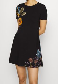 Czarna sukienka z krótkimi rękawami, ozdobiona kolorową haftem kwiatowym z jednej strony i w luźnym kroju, wykonana z miękkiego materiału jersey.