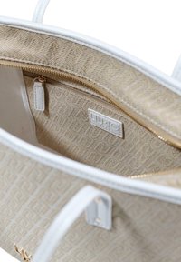 Beige tygväska inuti med en ficka med dragkedja, guldfärgad dragkedja och präglad logoplåt. Vita läderdetaljer och mjuk textur.