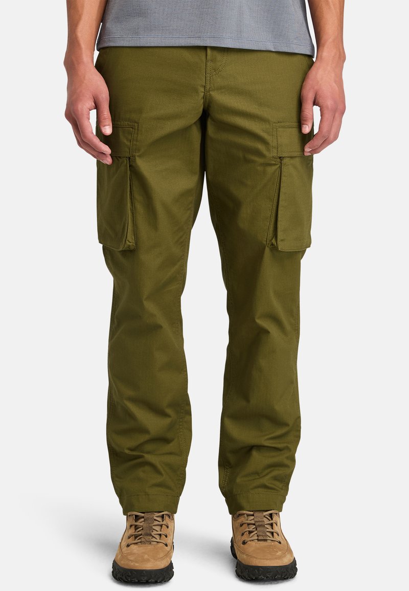 Pantaloni cargo verde oliva con due grandi tasche laterali, design a gamba dritta, tessuto resistente, indossati con stivali da escursione marroni.