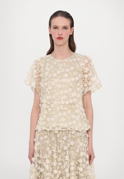 Haut beige à broderies florales avec des manches courtes, présentant un design superposé et une texture transparente, orné de détails floraux en trois dimensions.