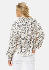 Beige blouse with navy heart pattern, long sleeves, buttoned cuffs, and a relaxed fit, worn over white pants. Texture appears smooth.