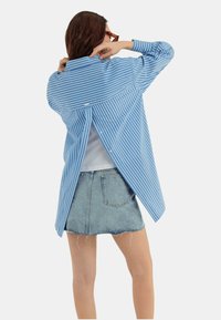 Camicia button-up a righe blu e bianche con vestibilità rilassata, dotata di una fessura sul retro e maniche lunghe, abbinata a una mini gonna in denim.