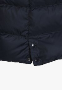 Tkanina námořnické modré péřové bundy má prošívaný vzor, hladký povrch a detail s patentním knoflíkem v dolním levém rohu.