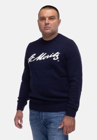 Maglione lavorato a maglia blu navy con logo bianco "St. Moritz", scollatura rotonda, polsini e orlo a coste, indossato con jeans azzurri chiari.