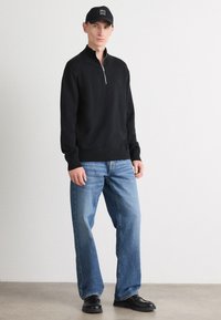 Czarny sweter na zamek, niebieskie dżinsy i czarne buty. Sweter ma długie rękawy i wysoki kołnierz. Czarna czapka z białym napisem dopełnia look.