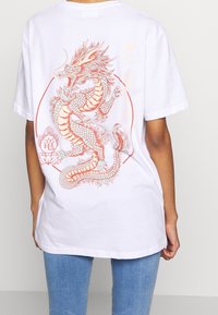 Bílý bavlněný tričko s velkým červeným a modrým drakem na zadní straně, s propracovanými detaily šupin a květinovými akcenty.