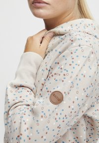 Beige hettegenser med flerfarget prikkemønster i blått, brunt og hvitt. Har ribbestrikkede mansjetter og et lærlogo-patch på armen.