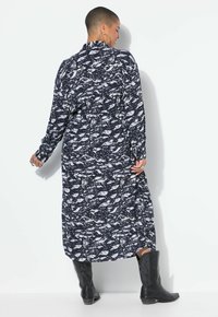 Vestido de manga larga y largo hasta el tobillo en color navy con un patrón abstracto blanco, que presenta un cuello con solapa y bolsillos laterales. Llevado con botas negras.