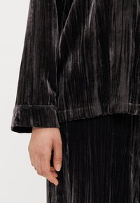 Samallinen loungewear-setti tummanharmaana, jossa on pystysuuntainen ribbitekstuuri. Löysä istuvuus ylhäällä, jossa on laskostetut hihansuut ja pehmeä, sileä kangas. Käsi näkyvissä.
