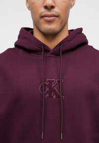 Hanorac maroniu cu şnururi, logo „CK” brodat în fir asortat şi un material moale, texturat.