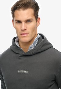 Sudadera gris con capucha de cordón, logo prominente de "SUPERDRY" en blanco, llevada sobre una camisa de rayas azul y blanca con cuello. Textura suave.
