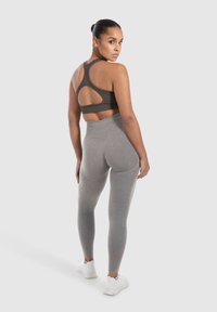 Bruine sport-bh met racerback-design en gaasdetails, gecombineerd met hooggesneden grijze legging. Beide zijn gemaakt van rekbaar materiaal. Witte sneakers.
