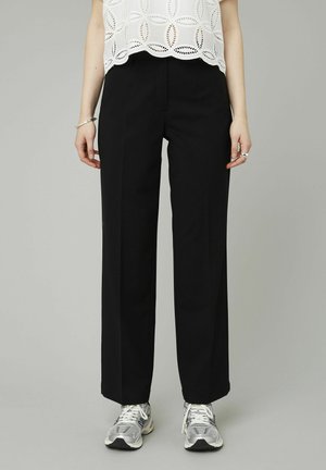 Pantalons larges noirs en tissu structuré, avec une finition lisse et des plis à l'avant, associés à un chemisier blanc à découpes et des baskets.