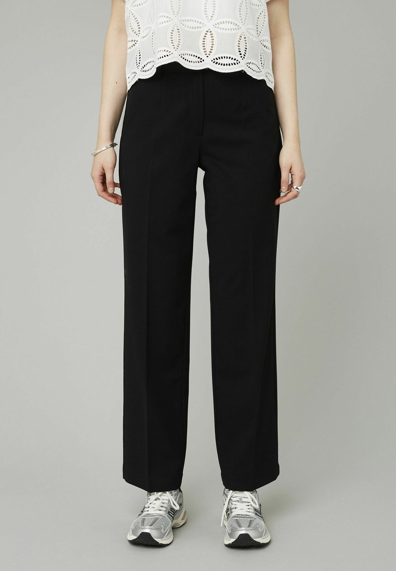 Pantalons larges noirs en tissu structuré, avec une finition lisse et des plis à l'avant, associés à un chemisier blanc à découpes et des baskets.