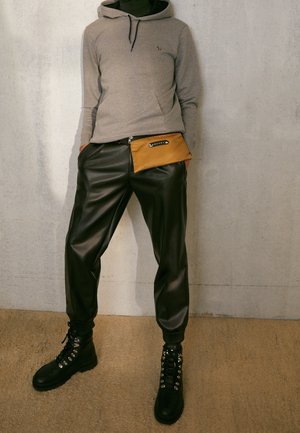 Personne portant un sweat à capuche gris, un pantalon de jogging noir en similicuir, des bottes noires à lacets et une pochette marron à la taille, debout contre un mur en béton.