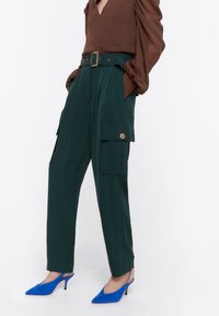 Zelené высокé cargo kalhoty s plisováním, bočními kapsami a širokým páskem. Slučovány s hnědou blůzou a modrými botami s špičatou špičkou.