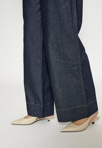 Džínové nohavice s širokými nohavicami s kontrastným bielym prešívaním, kombinované so svetlo béžovými slingback opätkami s ostrou špičkou, predstavujúce čistý, minimalistický dizajn.