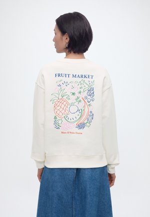 Mujer con el cabello corto y oscuro, vistiendo una sudadera blanca con ilustraciones coloridas de frutas y el texto "FRUIT MARKET" en la espalda, combinada con una falda vaquera.