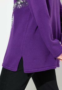 Maglione lavorato a maglia viola con uno spacco laterale, caratterizzato da un design di paillettes argento sulla parte anteriore, abbinato a pantaloni neri.