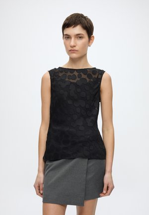 Femme aux cheveux courts portant un haut noir sans manches avec des motifs texturés et une mini-jupe asymétrique grise, debout devant un fond uni.