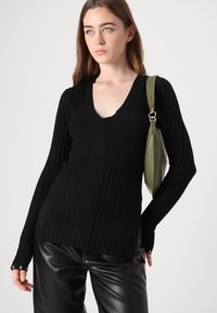 Pull noir à côtes avec un col en V et des manches allongées. Porté avec un pantalon en cuir noir brillant et portant un sac vert.