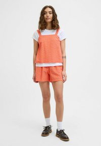 Žiarivo oranžový kockovaný crop top so širokými ramienkami na bielom tričku a zodpovedajúcich šortkách. Masívne čierne topánky so šnúrkami a biele ponožky.