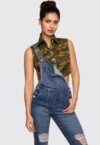 Camo-patterned, sleeveless shirt with a collared neck paired with blue denim overalls featuring distressed details and adjustable straps.