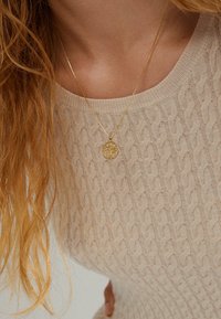 Collana di pendente d'oro con un design ad albero, indossata su un maglione a maglia beige chiaro. La sottile catena mette in risalto i dettagli intricati del pendente.