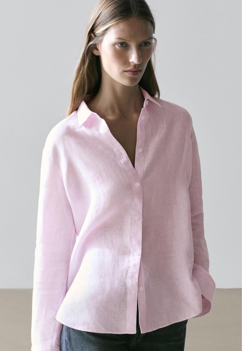 Frau trägt ein hellrosa langärmliges Hemd mit Knöpfen und Kragen, steht vor einem schlichten hellgrauen Hintergrund.