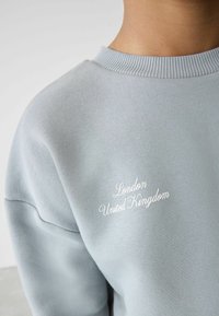 Lys blå sweatshirt med ribbet krave og subtil tekstur. Har hvid broderet tekst, der står "London United Kingdom" på forsiden.