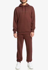Bruine katoenen hoodie en bijpassende joggers. De hoodie heeft een zak aan de voorkant en trekkoorden, met subtiele branding op beide stukken.