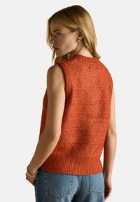 Õlavarteta kudumispullover põletatud oranžis värvitoonis, millel on tekstureeritud rombimuster ja soonik serv, sobitatud helesiniste teksadega.