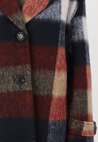 Manteau en mélange de laine à carreaux, avec des blocs rouge, bleu marine et beige. Des boutons noirs et des poignets repliés ajoutent du détail au tissu texturé.