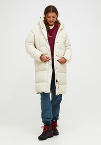 Pufferjack in crèmekleur met een capuchon, voorzien van grote, gewatteerde secties, drukknoopsluitingen en zijzakken. Gepaard met blauwe sweatpants en zwarte laarzen.