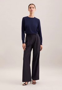 Maglia a maniche lunghe color navy abbinata a pantaloni neri a vita alta e a gamba larga. Il modello indossa décolleté nere con cinturini. Tessuto liscio, vestibilità su misura.