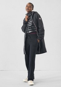 Donna con un gilet imbottito nero indossato sotto un lungo giubbotto nero con cappuccio, pantaloni neri e sneakers bianche, in piedi contro un muro chiaro e semplice.