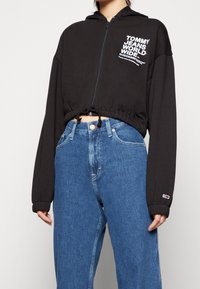 Sudadera negra recortada con cierre de cremallera y dobladillo con cordón, con texto de logo blanco en el pecho y puños de canalé. Combinada con jeans azules.