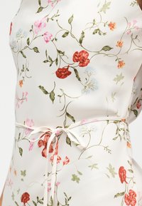 Λευκό ύφασμα με κόκκινο, ροζ και πορτοκαλί floral σχέδιο, πράσινα φύλλα και λεπτή λευκή ζώνη δέσιμος γύρω από τη μέση.