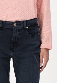 Jeans de mezclilla oscura con cintura alta, bolsillo delantero y detalle de botón dorado. Combinados con una blusa de manga larga con rayas naranjas.
