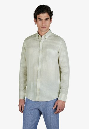 Homme portant une chemise à rayures vert clair avec une poche poitrine et un pantalon bleu clair, debout sur un fond blanc.
