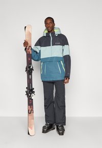 Snowboarder com uma jaqueta verde-água, azul escuro e verde limão, calças pretas e botas de esqui, segurando esquis com ligações ajustáveis.