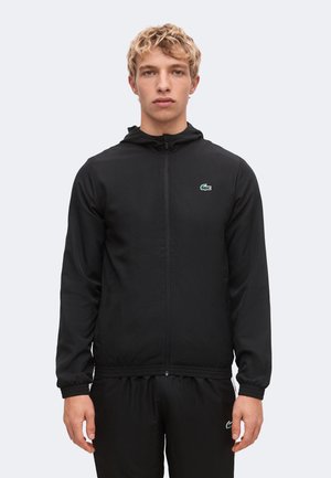 Junger Mann mit blondem lockigem Haar, der eine schwarze Kapuzenjacke mit Reißverschluss und schwarze Hose mit grünem Krokodil-Logo trägt.