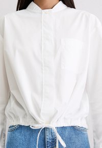 White, long-sleeve button-up shirt with a band collar, drawstring waist, and a single breast pocket. Worn over blue denim.