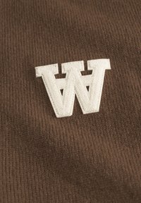 Brązowa żebrowana tkanina z dużą białą haftowaną literą "W" umieszczoną centralnie. Faktura jest miękka, jednolita i ciasno tkana.