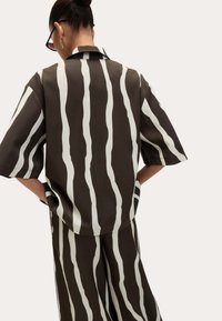 Camicia marrone con strisce bianche ondulate, maniche corte e colletto. Abbinata a pantaloni coordinati, che presentano un tessuto fluido e leggero.