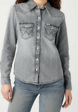 Osoba ubrana w wyblakłą szarą jeansową koszulę zapinaną na guziki z klapkowymi kieszeniami na piersi oraz niebieskie dżinsy, widoczna od szyi do bioder.