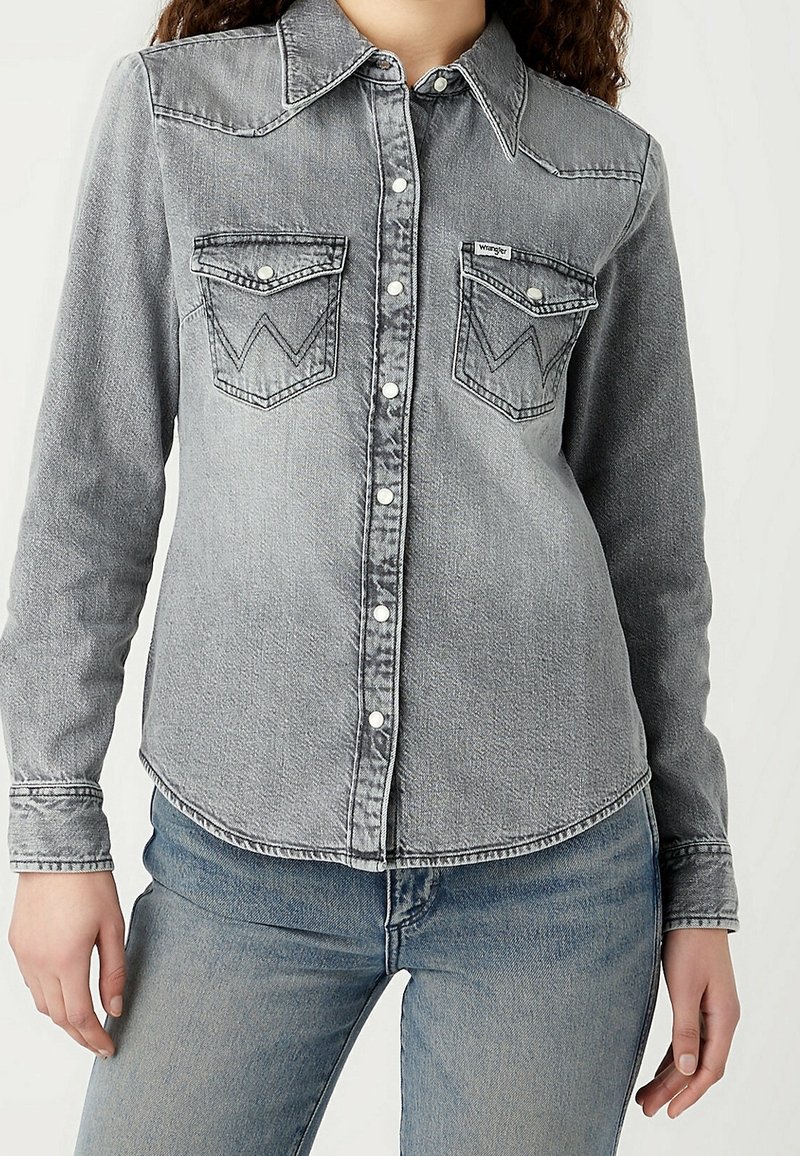 Personne portant une chemise en jean gris délavé à boutons avec poches poitrine à rabat et un jean bleu, visible de la nuque jusqu'aux hanches.